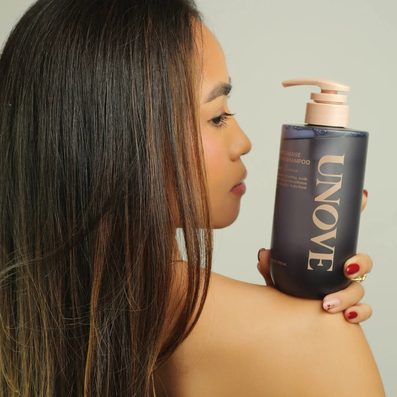 Woman with long healthy hair holding UNOVE shampoo bottle against neutral background