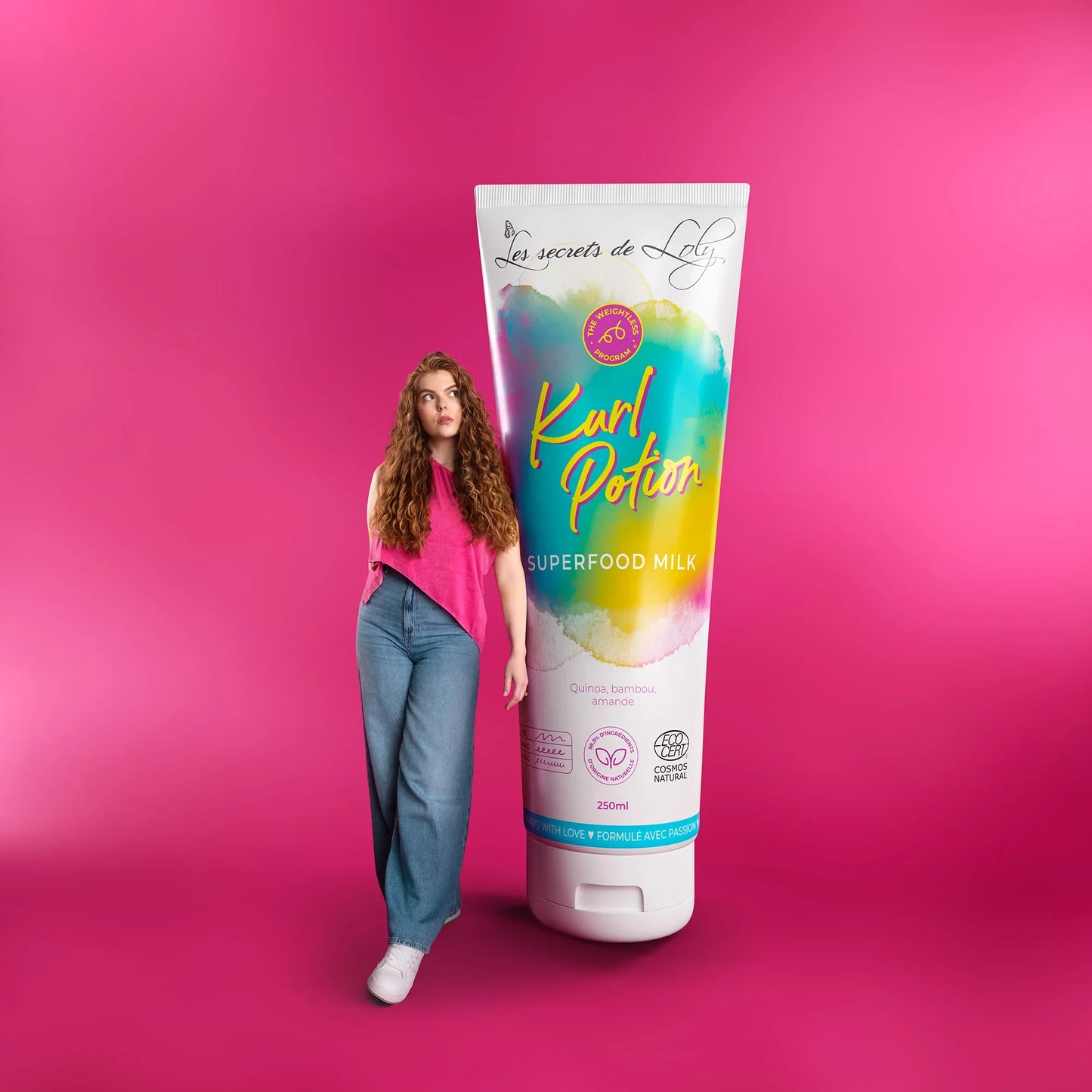 Woman with long curly hair standing beside large Kurl Potion Superfood Milk haircare tube on pink background