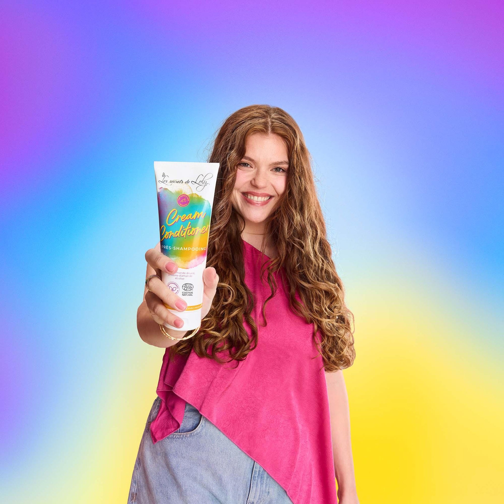 Smiling woman with long curly hair holding cream conditioner tube on colorful gradient background