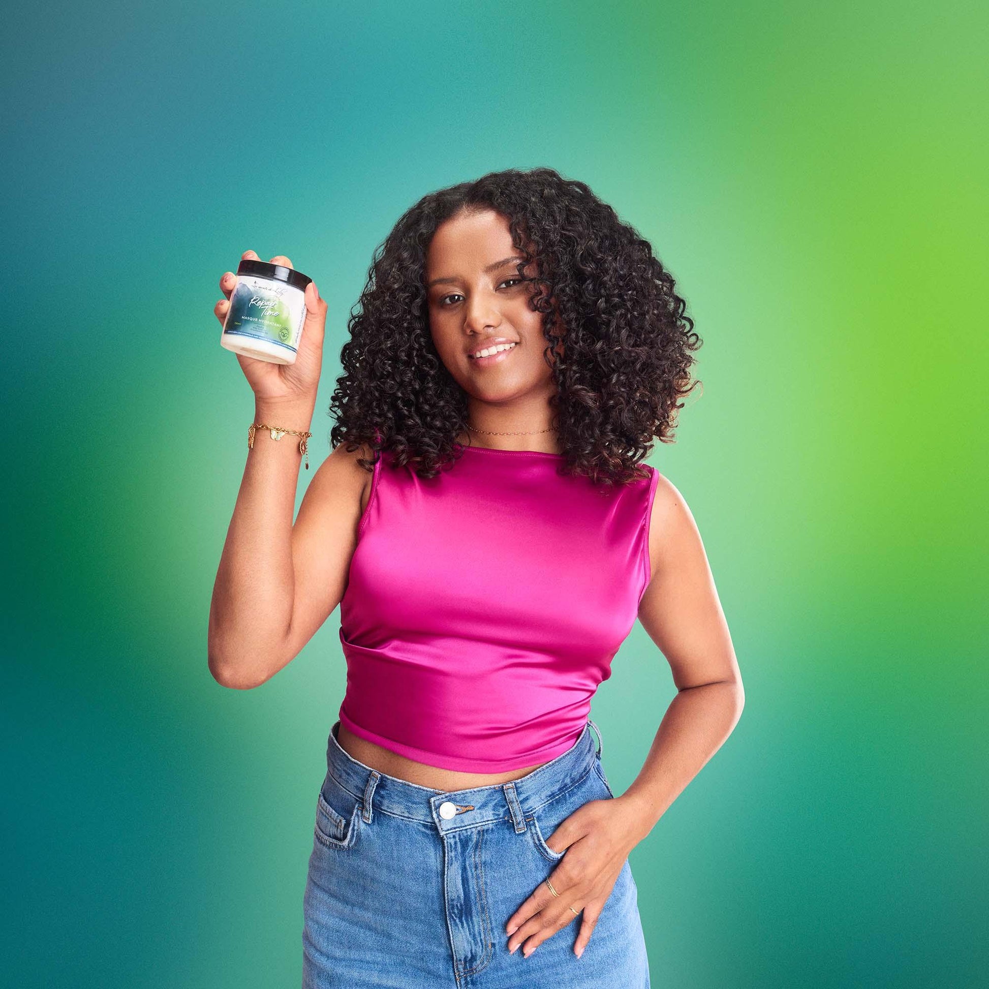 Woman with curly hair in pink top holding haircare product against green gradient background.