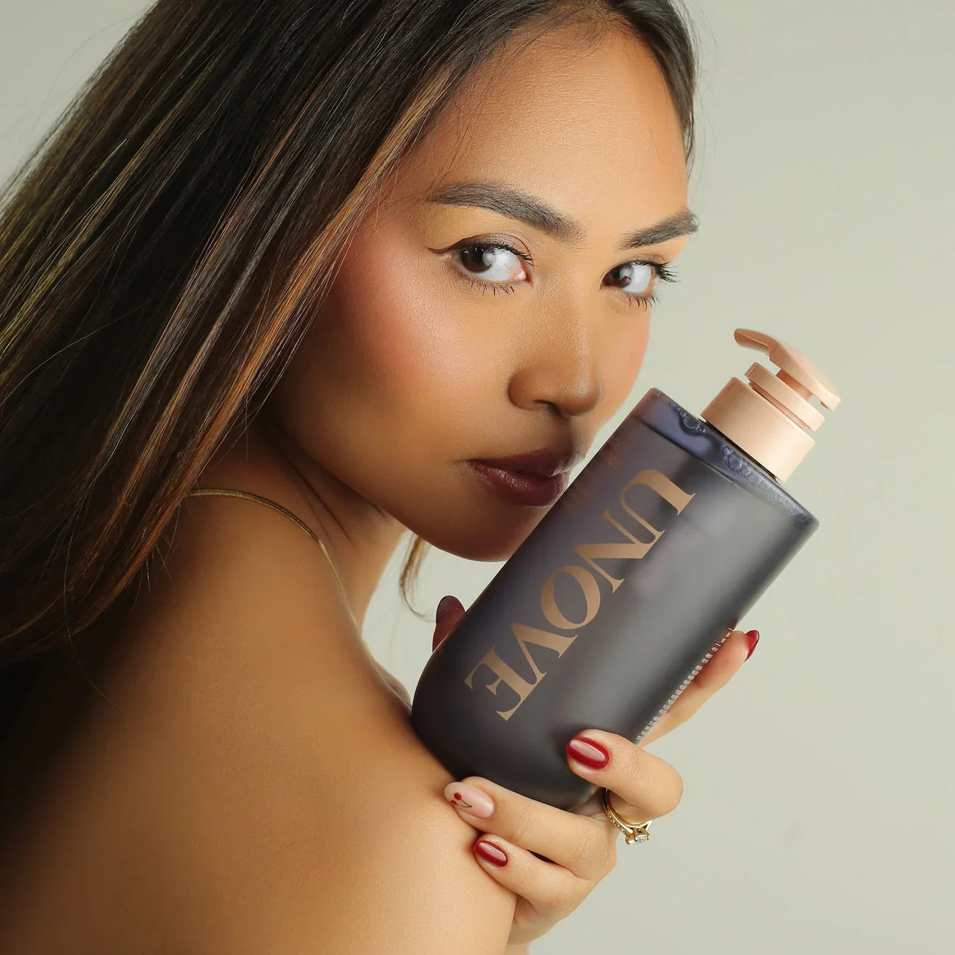 Woman with long hair holding UNOVE haircare bottle close to face, neutral background
