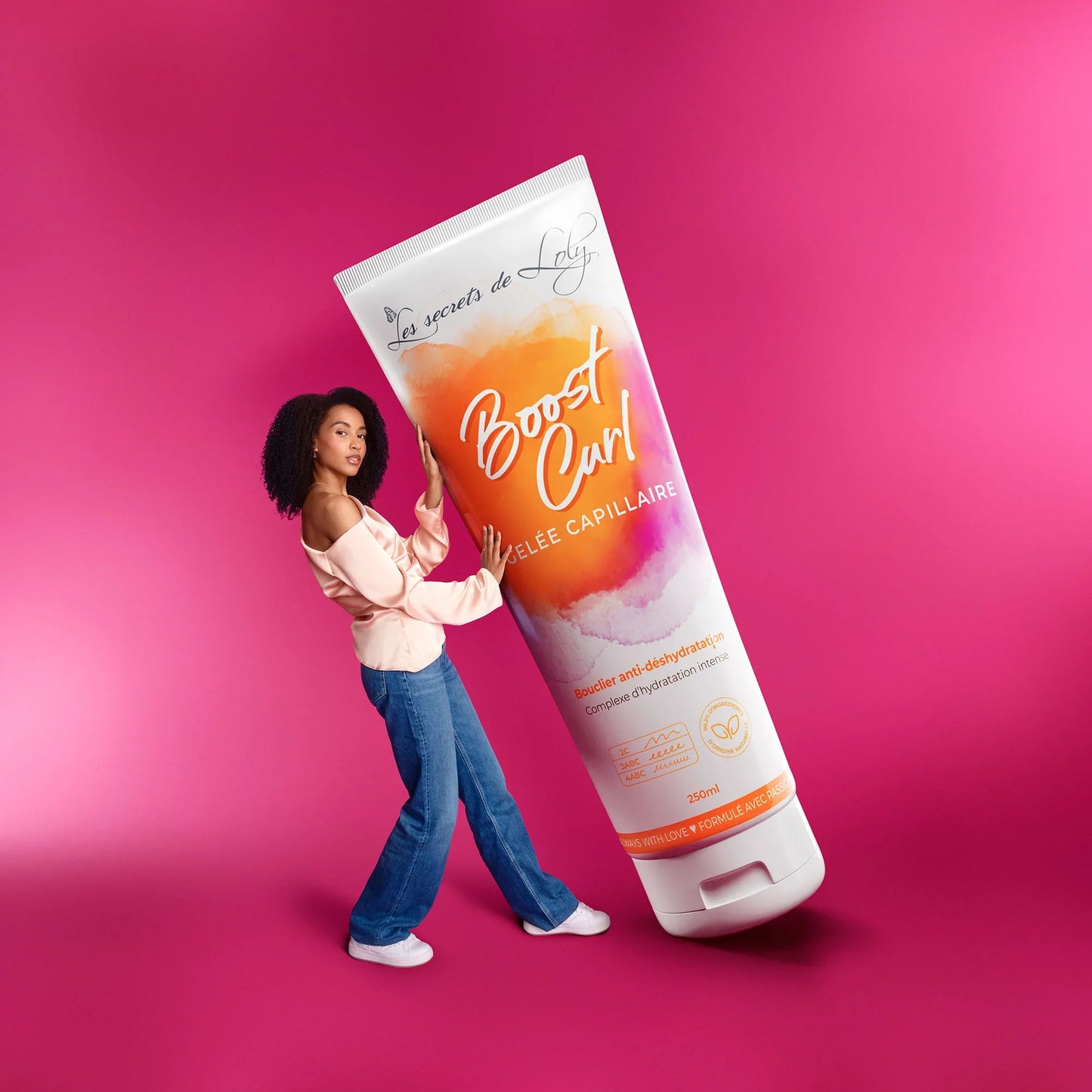 Woman with curly hair poses beside oversized Boost Curl hair gel tube on pink background