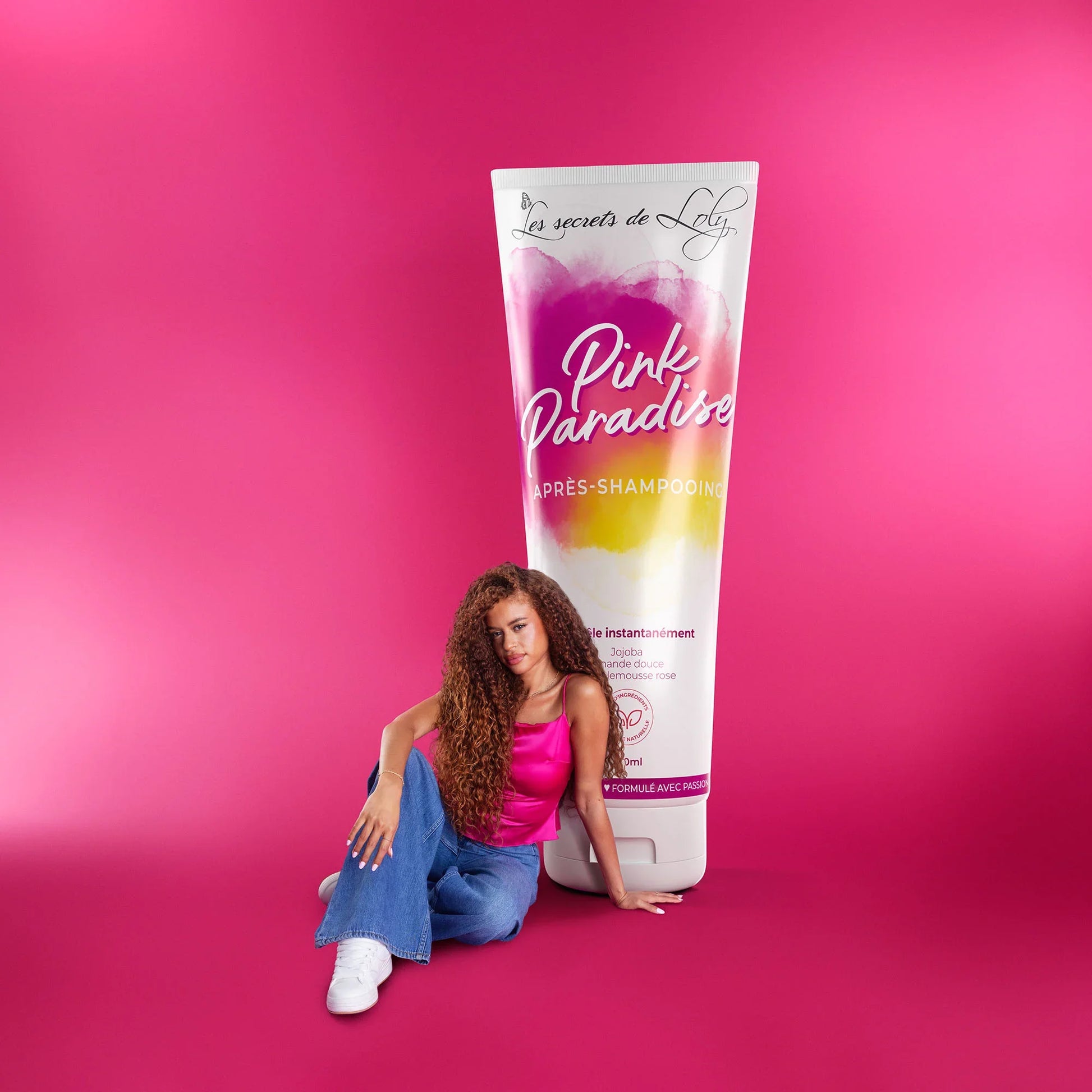 Woman with curly hair sitting beside oversized Pink Paradise hair care product tube on pink background