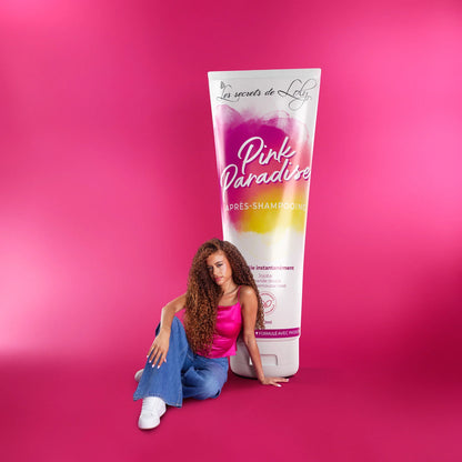 Woman with curly hair sitting beside oversized Pink Paradise hair care product tube on pink background