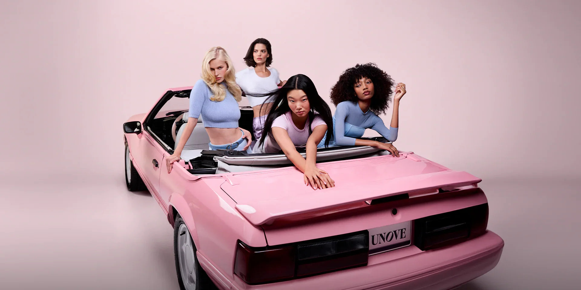 Four women with diverse hair types posing in a pink convertible, soft pink background